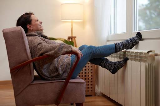 Old woman sitting in chair in front of radiator with her feet warming up on it.
